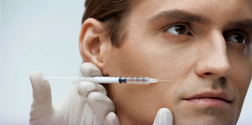 The young man who looks elsewhere and the doctor's hand holds the glove of his hand and holds the person with one hand and with the other is injecting fat with a syringe - injecting fat into men's face.