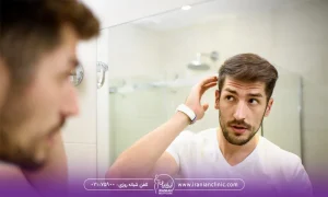 تصویر مربوط به یک آقاست که تیشرت سفید پوشیده و جلوی آینه به خودش نگاه می کنه و به موهاش دست می کشد