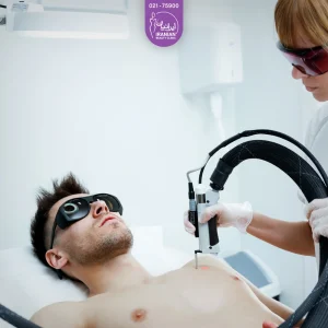 عکس مرد جوان که روی تخت خوابیده و عینک به چشم دارد و توسط اپراتور دستگاه لیزر بدن او لیزر می شود - لیزر موهای زائد مردان